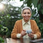 Elderly woman sitting in her garden with a cup of coffee and a laptop, bathed in early morning sunlight, symbolizing new beginnings in online business