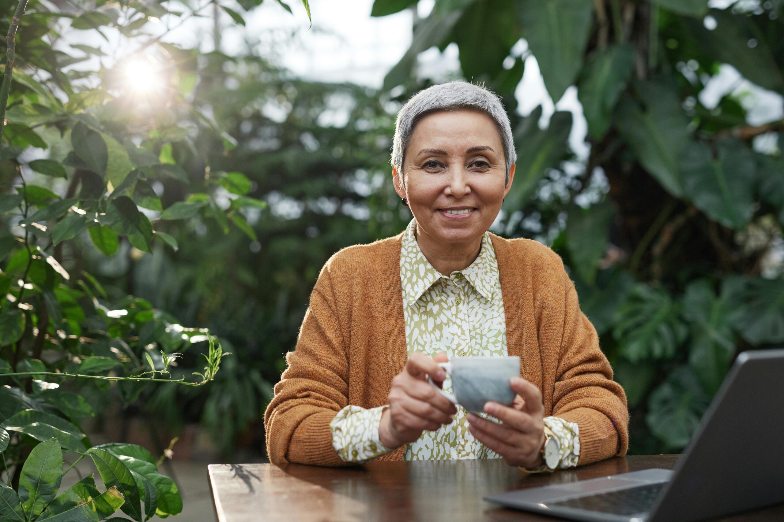 Elderly woman sitting in her garden with a cup of coffee and a laptop, bathed in early morning sunlight, symbolizing new beginnings in online business