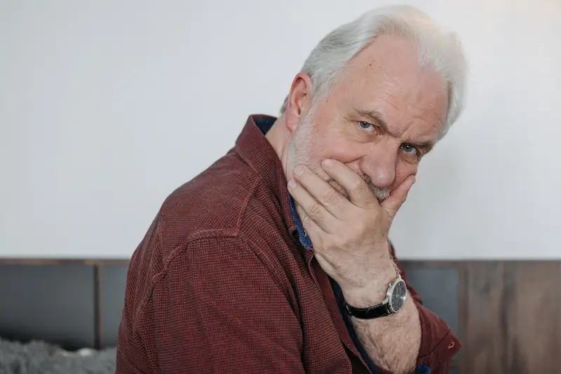 Close-up portrait of a thoughtful elderly man with white hair wearing a red shirt, hand on face.